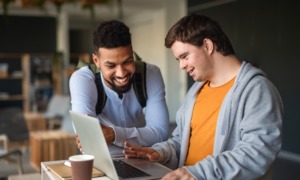 Disabled student college & job transition, disability inclusion: Teen boy with Down syndrome sits working on laptop next to young Black man tutoring him