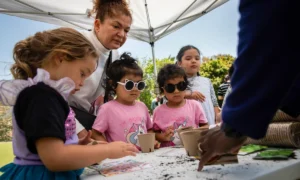 Head Start funding: Several young, ethnically mixed children sit at table doing crafts while woman stands looking over heir shoulders.